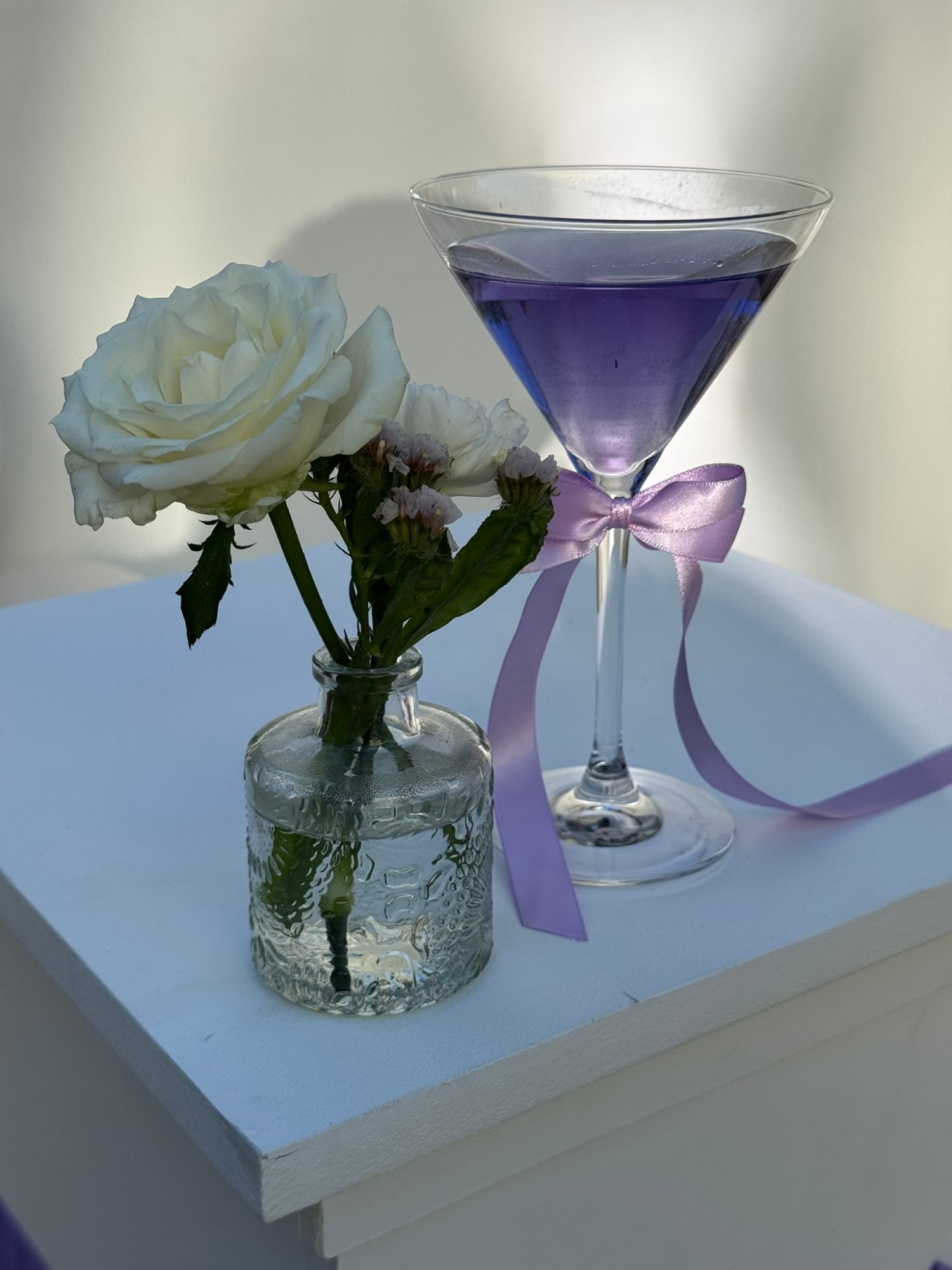 Cocktail violet dans un verre à martini noué d'un ruban lilas, à côté d'une rose blanche en soliflore