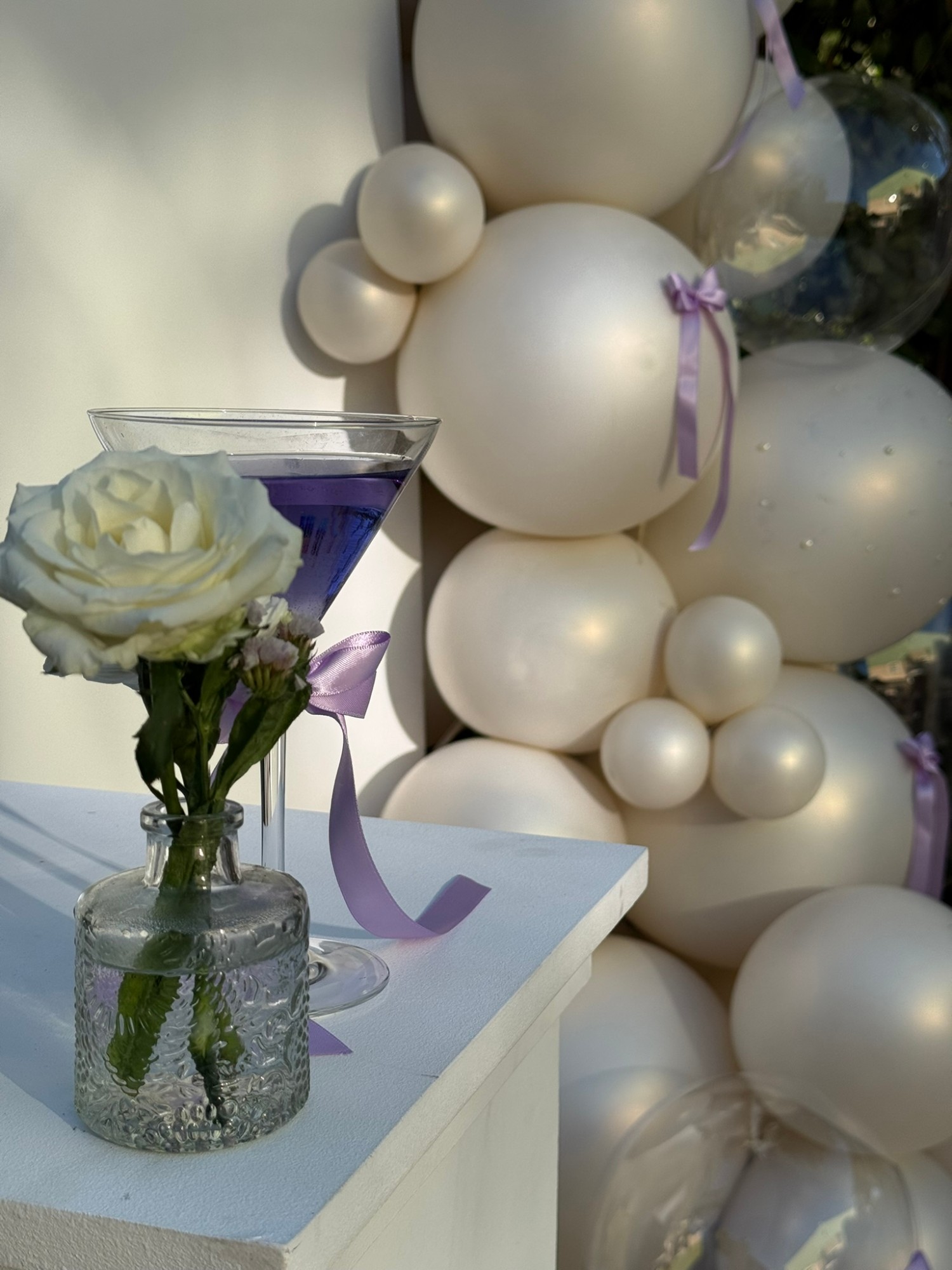 Détail : verre à cocktail violet et rose blanche devant la cascade de ballons nacrés aux nœuds lilas