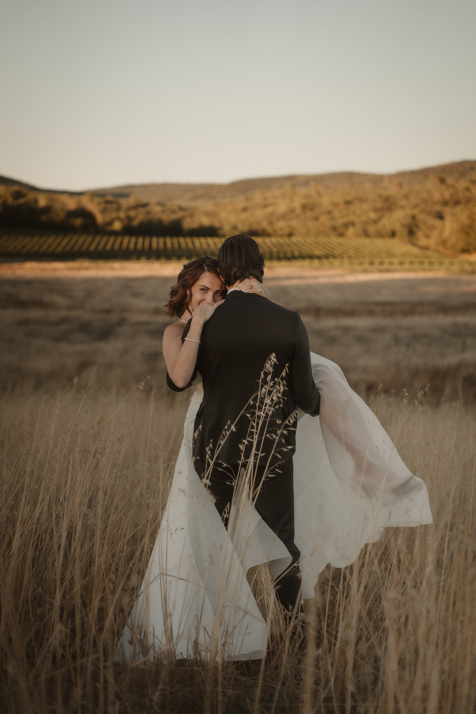 Le marié porte la mariée dans les hautes herbes, robe au vent, shooting Vendanges Chic