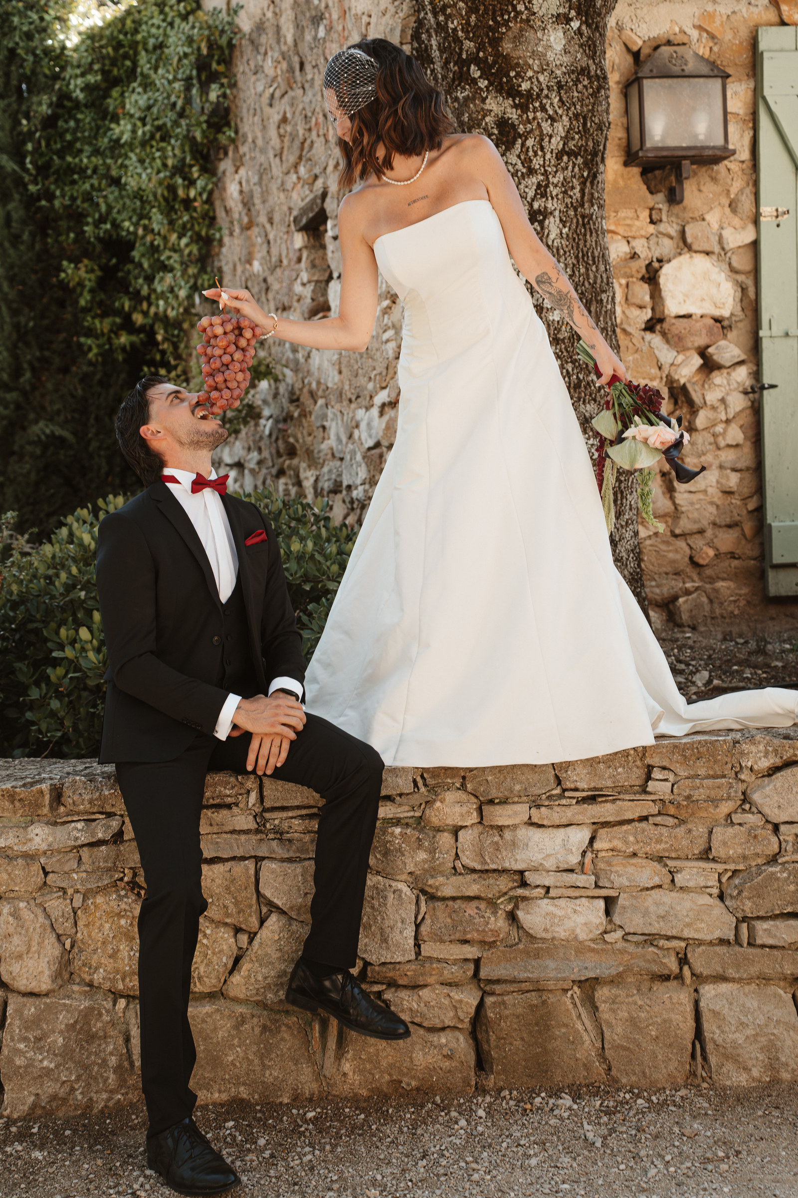 Sur le muret en pierre, la mariée offre une grappe de raisin à son mari — shooting Vendanges Chic aux Terres Saint-Hilaire