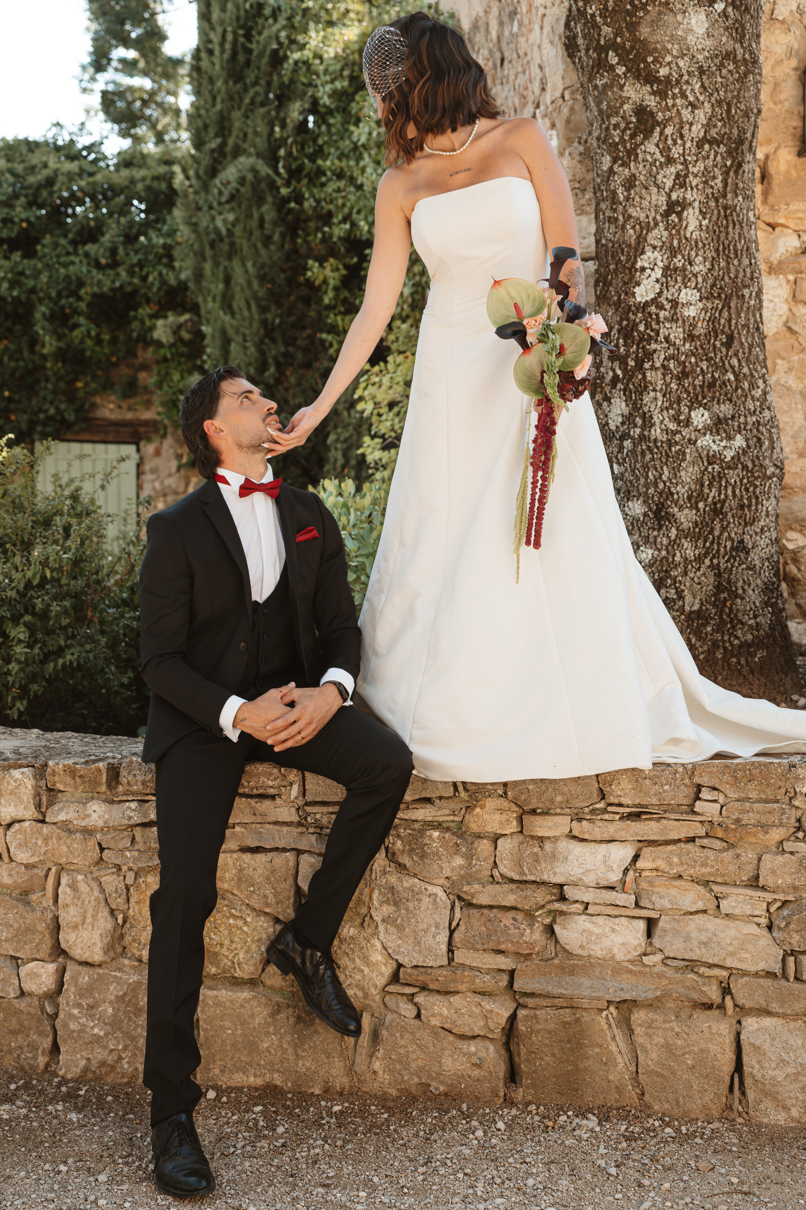 Sur le muret en pierre, la mariée tient tendrement le visage de son mari, Les Terres Saint-Hilaire